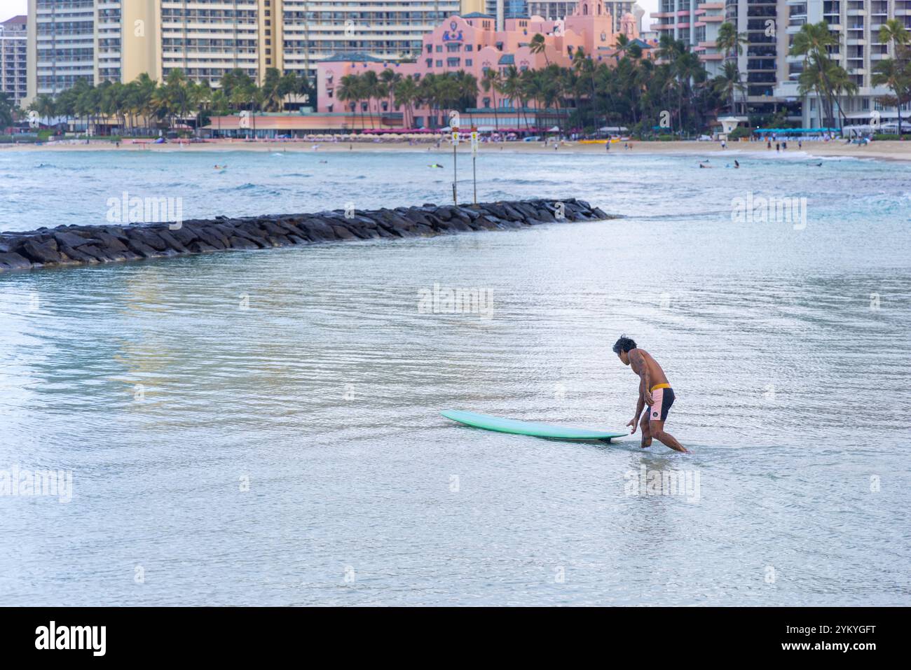 Un uomo è in piedi in acqua con una tavola da surf verde. Sta indossando un costume da bagno Foto Stock