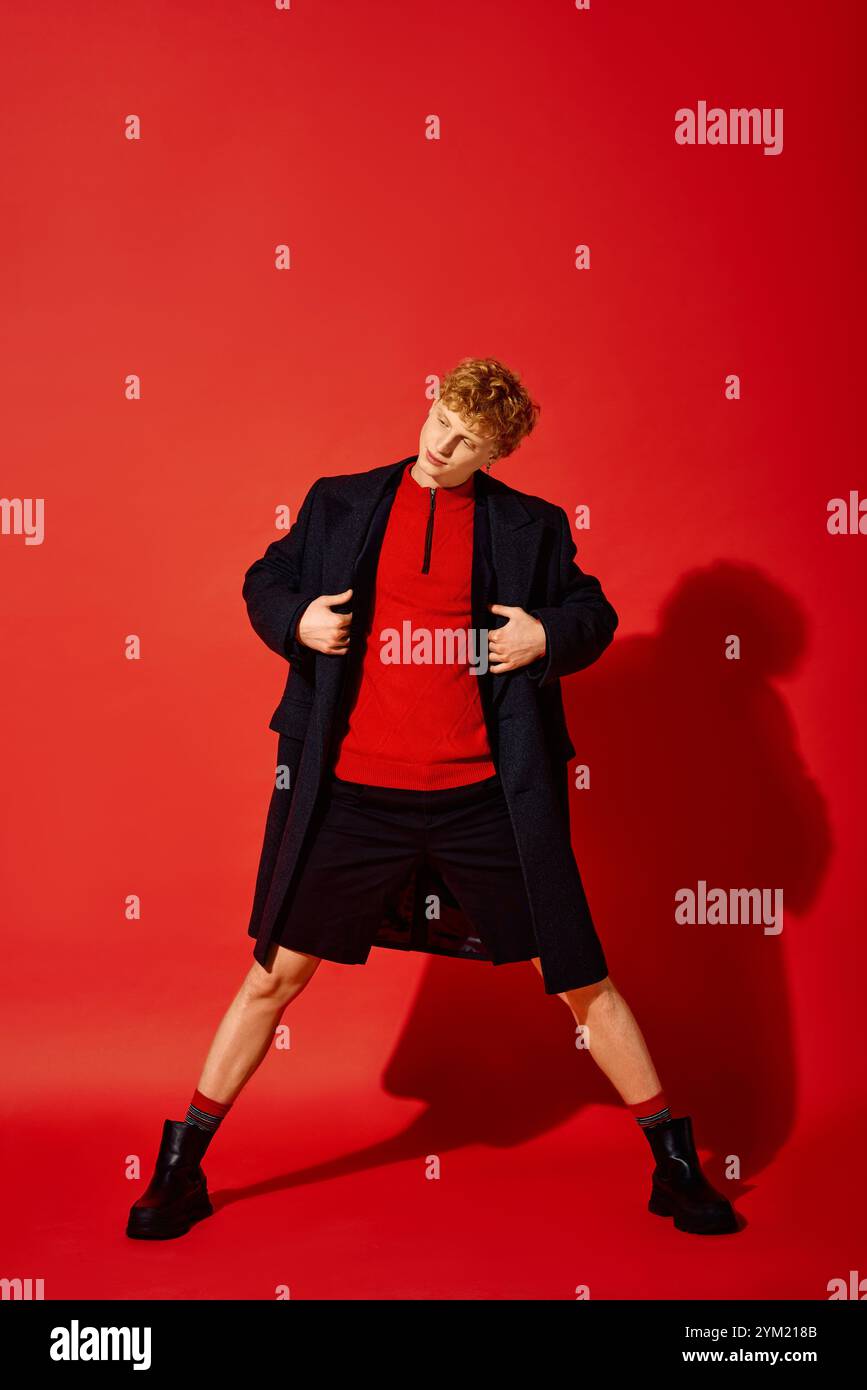 Un uomo di grande impatto mette in mostra la moda natalizia moderna su un vivace sfondo rosso. Indossa con sicurezza un elegante top rosso abbinato a un cappotto elegante, embo Foto Stock