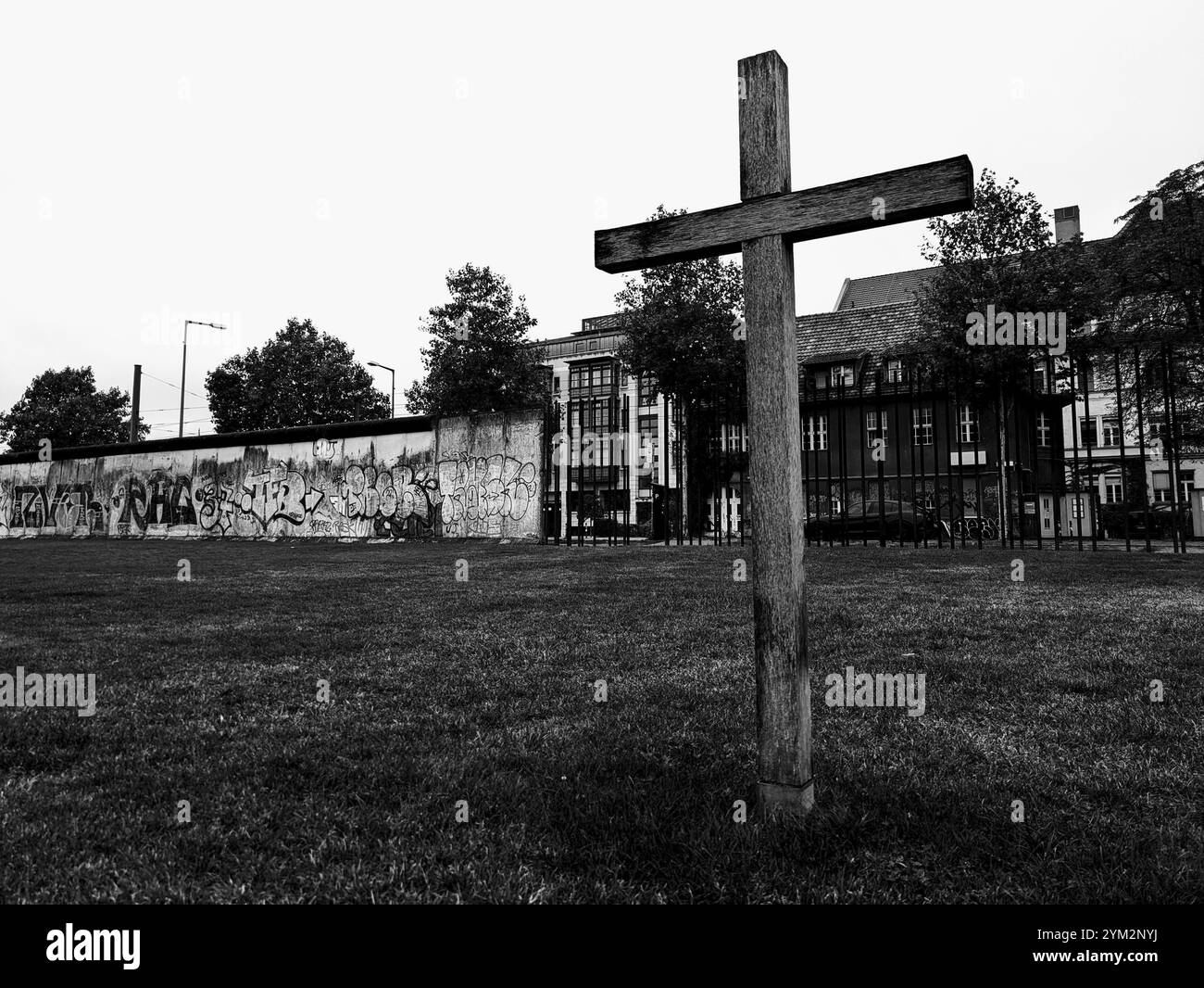 Immagine in bianco e nero di una croce di legno vicino al muro di Berlino ricoperta di graffiti, che evoca un'atmosfera cupa, Una croce in piedi di fronte a un graffito Foto Stock