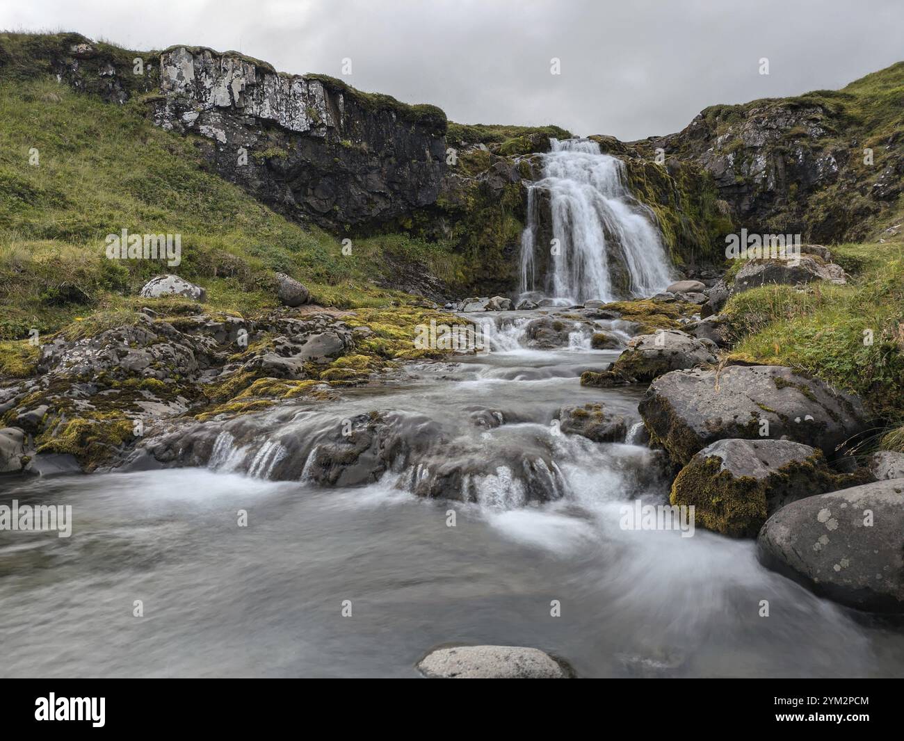 Cascata che si estende su rocce muschiate con acqua corrente e vegetazione sotto un cielo nuvoloso. Islanda Foto Stock