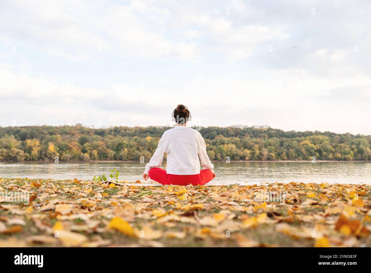 Donna con vista posteriore che medita all'aperto in riva al fiume in autunno, pratica yoga e trova armonia con la natura. Foto Stock