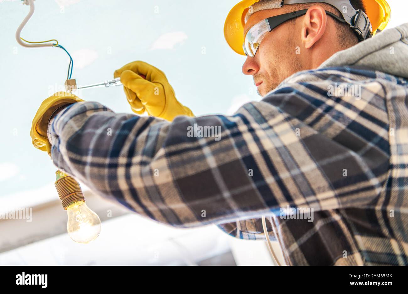 Un lavoratore esperto collega un apparecchio di illuminazione in un edificio, indossando un casco e guanti per la sicurezza. Impegno mirato a garantire una corretta installazione. Foto Stock