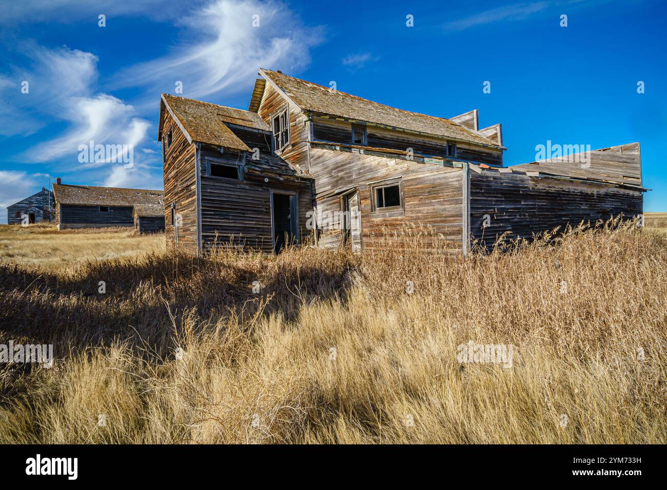 Una grande e vecchia casa si trova in un campo di erba alta. La casa è circondata da altri vecchi edifici, e il cielo è limpido e blu. La scena è tranquilla e.. Foto Stock