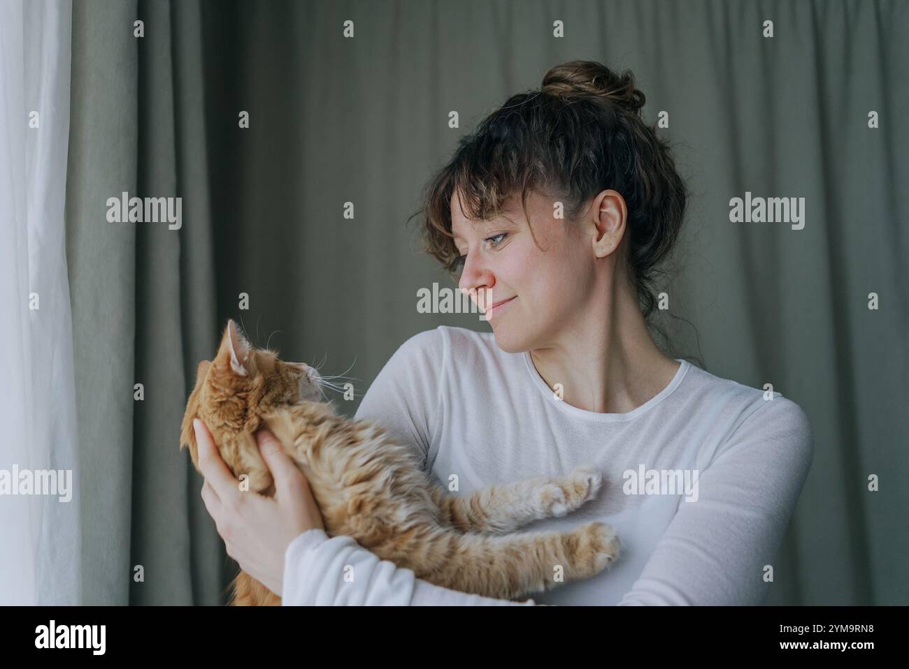 Giovane donna con i capelli scuri legati in un panino, che indossa una camicia bianca a maniche lunghe, tiene delicatamente un gatto di zenzero vicino a una finestra con tende verdi chiaro, creando una tranquilla scena domestica Foto Stock