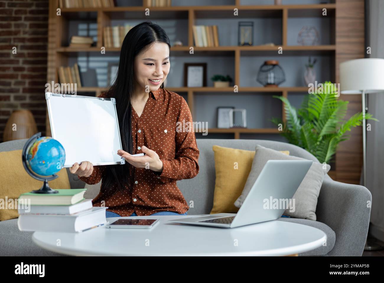 Donna asiatica che conduce una lezione online con laptop e lavagna. Interazione coinvolgente attraverso il supporto digitale, integrando concetti globali ed educativi da casa. Foto Stock
