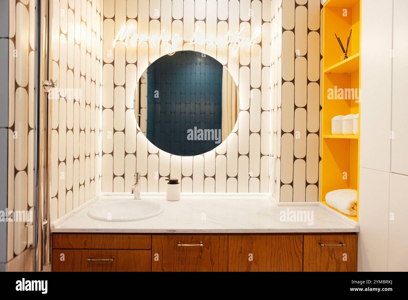 Interni del bagno moderni ed eleganti. Primo piano del piano di lavoro in marmo con lavandino e grande specchio illuminato rotondo. Accenti luminosi sugli scaffali gialli con Foto Stock