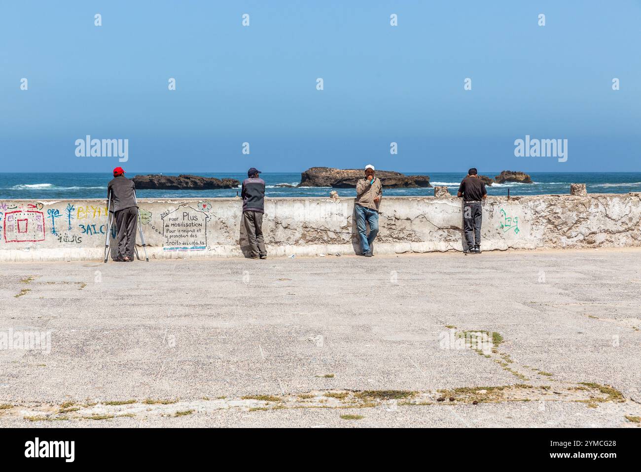 All'ingresso di Essaouira, appoggiato su una parete bassa vicino al mare, pochi uomini contemplarono il mare, i gabbiani volanti e i turisti che passano Foto Stock