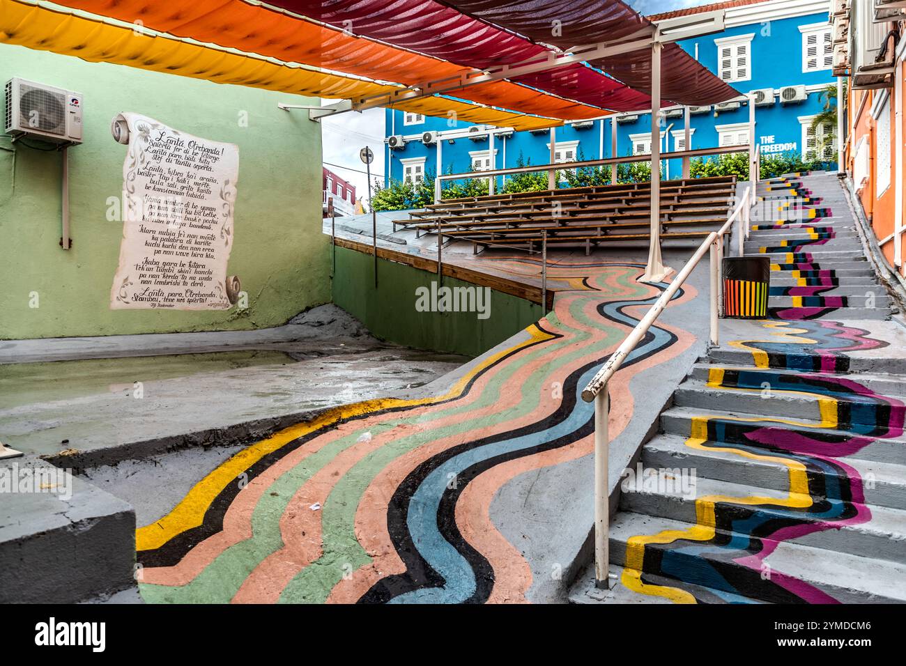 Il palco del festival e la tribuna all'aperto del KayaKaya Murals Festival si trovano nel quartiere di Otrabanda a Willemstad, Curaçao. Linee ondulate colorate sul terreno e scale, insieme a parasole in tessuto arancione, rosso e giallo, fanno parte dell'infrastruttura permanente di questo festival annuale di arte di strada, che trasforma ogni anno i vicoli del sito del patrimonio mondiale UNESCO con nuovi murales. A sinistra, sul muro, è dipinto un poema in papiamento. Foto Stock