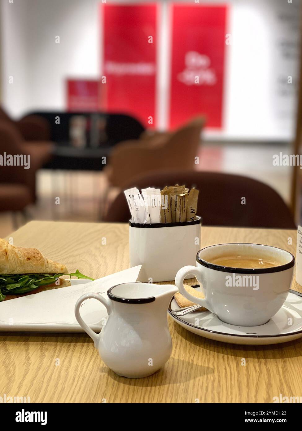 Una tazza da caffè bianca con impugnatura nera si trova su un tavolo accanto a una caraffa bianca. Un panino è su un tovagliolo sul tavolo Foto Stock