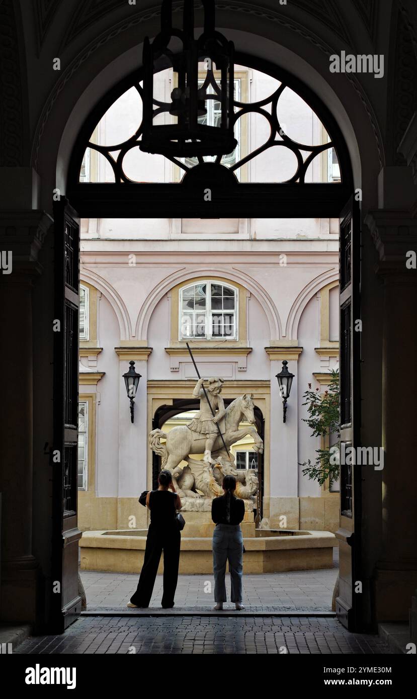 Due persone ammirano una fontana con una scultura di San Giorgio che uccide un drago in un cortile dello storico Palazzo dei Primati di Bratislava. Foto Stock