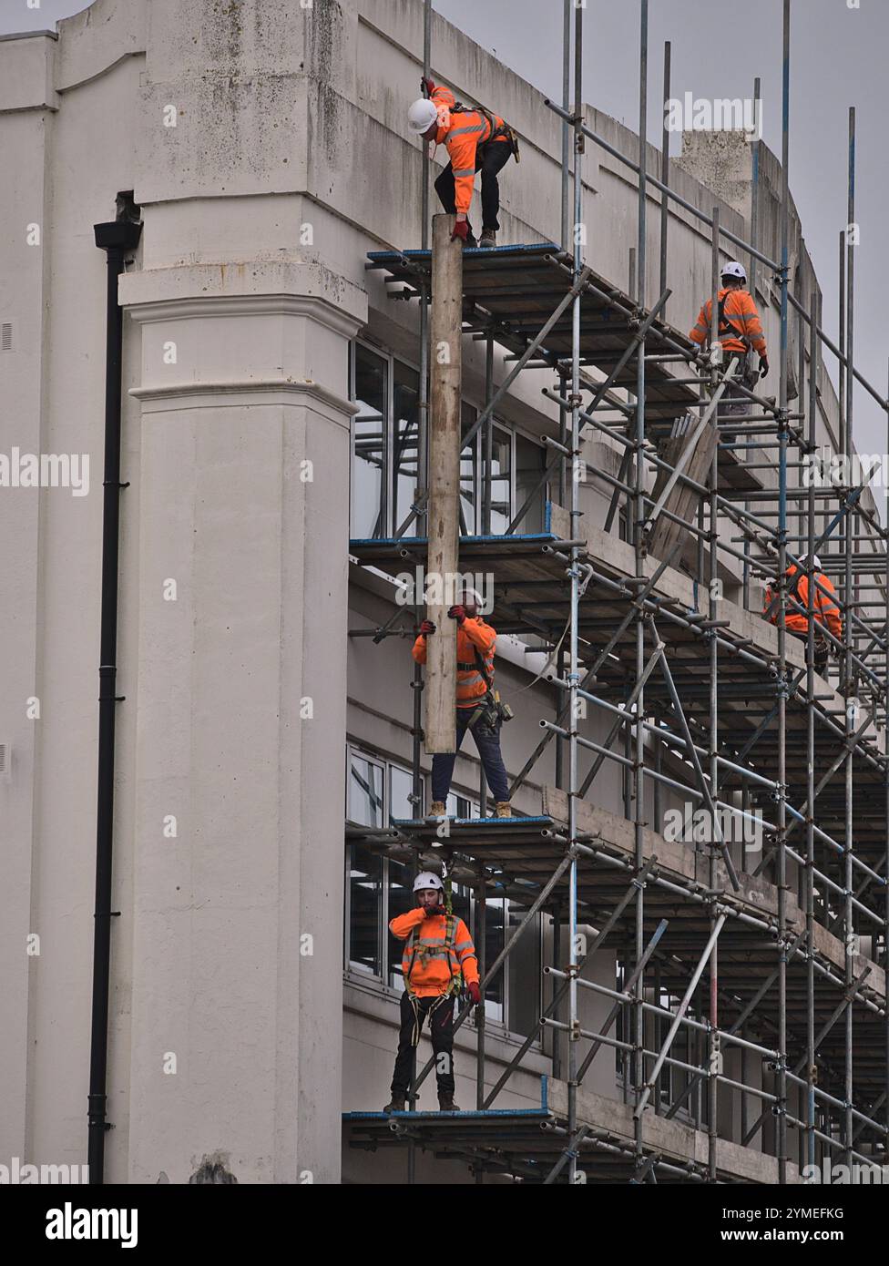 Ponteggio eretto da un team di ponteggi con cappelli arancioni ad alta visibilità e rigidi che lavorano insieme per sollevare le tavole dei ponteggi sulla piattaforma superiore Foto Stock