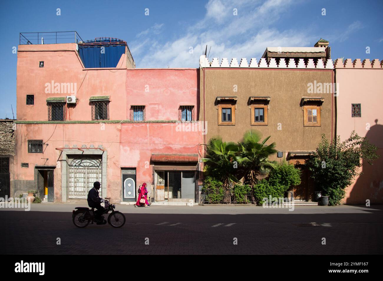 Colorata strada marocchina con edifici rosa e marrone, vegetazione vivace e una silhouette in moto contro un cielo blu limpido a Marrakech. Foto Stock