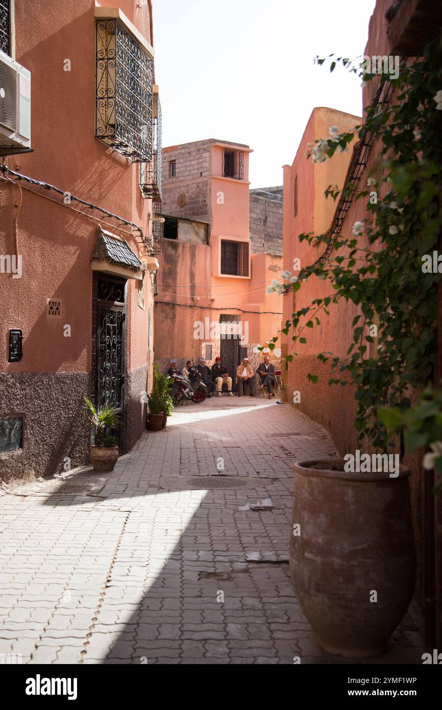 Tranquilla strada stretta nella medina di Marrakech con pareti in terracotta, piante in vaso e uomini locali seduti insieme al sole. Foto Stock