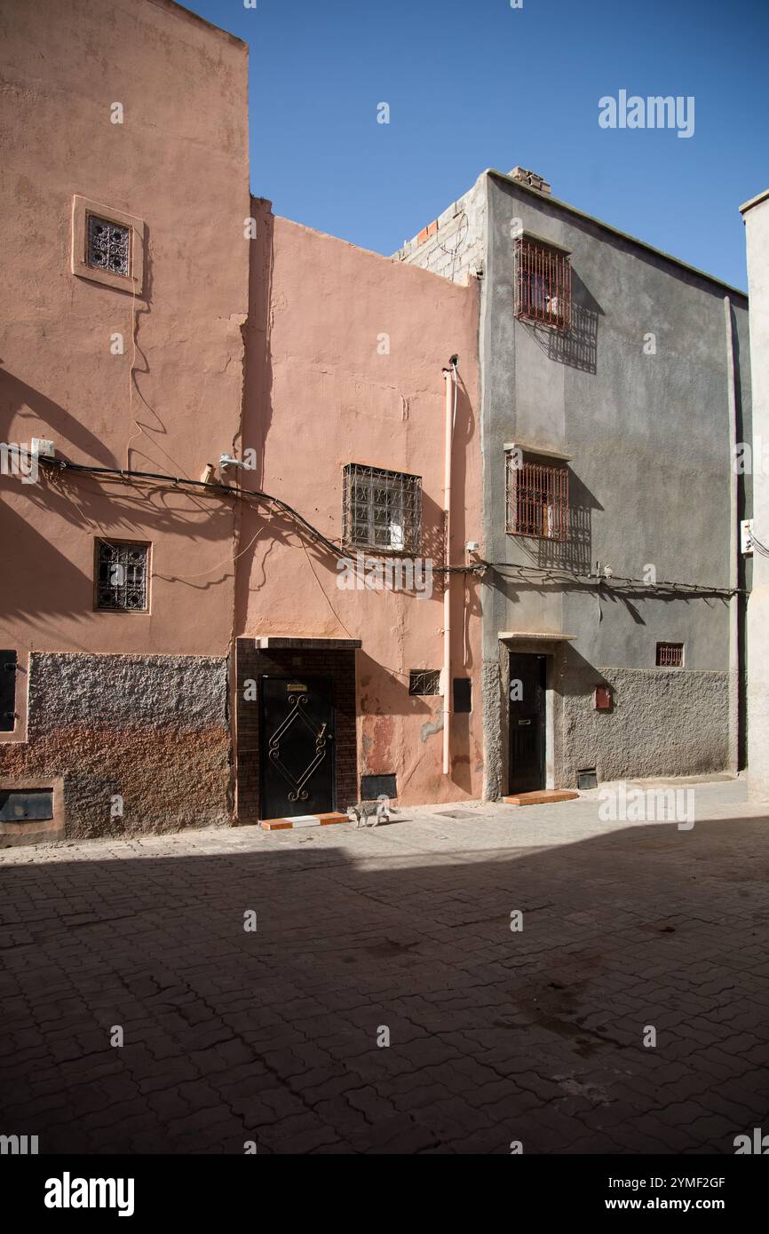 Rustica strada marocchina con edifici rosa e grigi, pareti testurizzate, griglie con finestre in ferro e una porta nera inondata di luce solare a Marrakech. Foto Stock