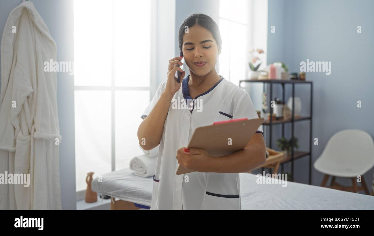 Donna nel centro benessere che indossa un'uniforme al telefono e tiene gli appunti in una tranquilla sala benessere. Foto Stock
