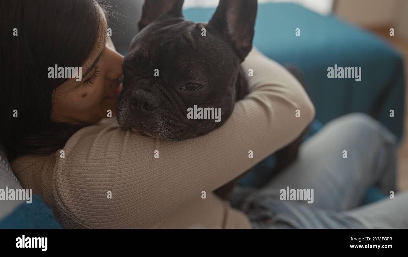 Una giovane donna ispanica coccola il suo bulldog francese nel salotto di casa sua, mostrando affetto e un ambiente interno accogliente con una luce delicata. Foto Stock