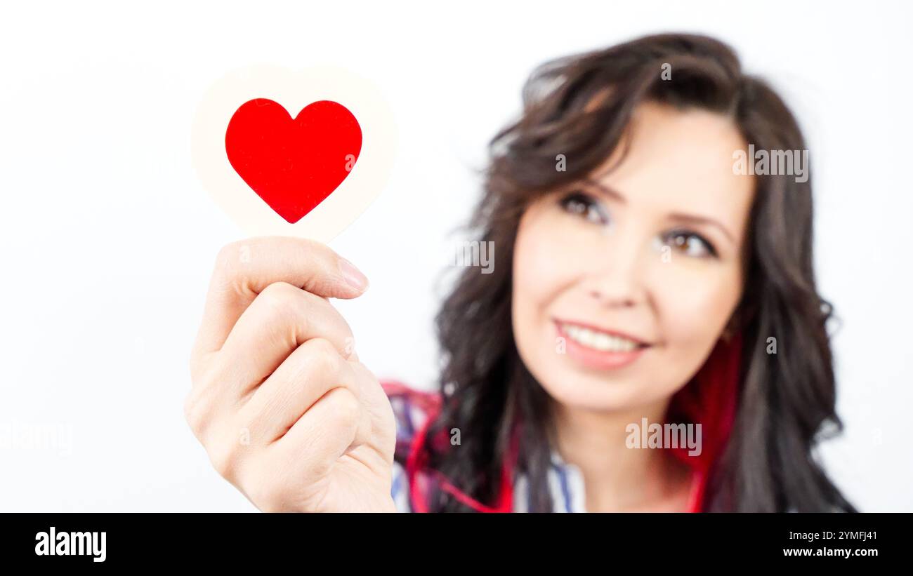 Giovane donna allegra con lunghi capelli scuri che tiene con gioia in mano un ritaglio rosso a forma di cuore. Foto Stock