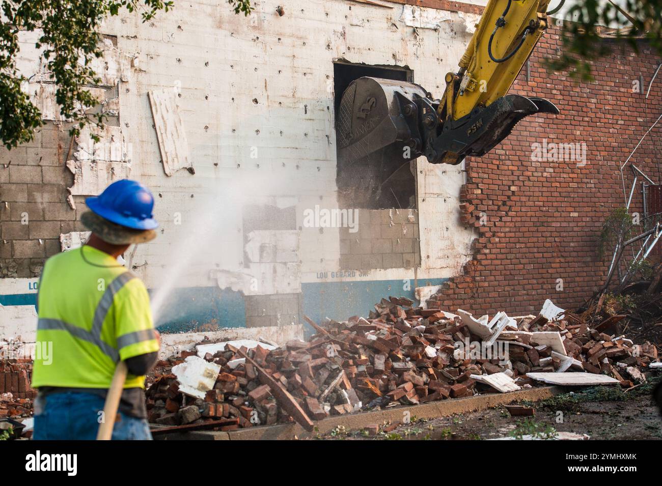 Demolizione in corso: I lavoratori gestiscono in modo sicuro la rimozione di una struttura in mattoni e calcestruzzo con macchinari pesanti e soppressione dell'acqua Foto Stock