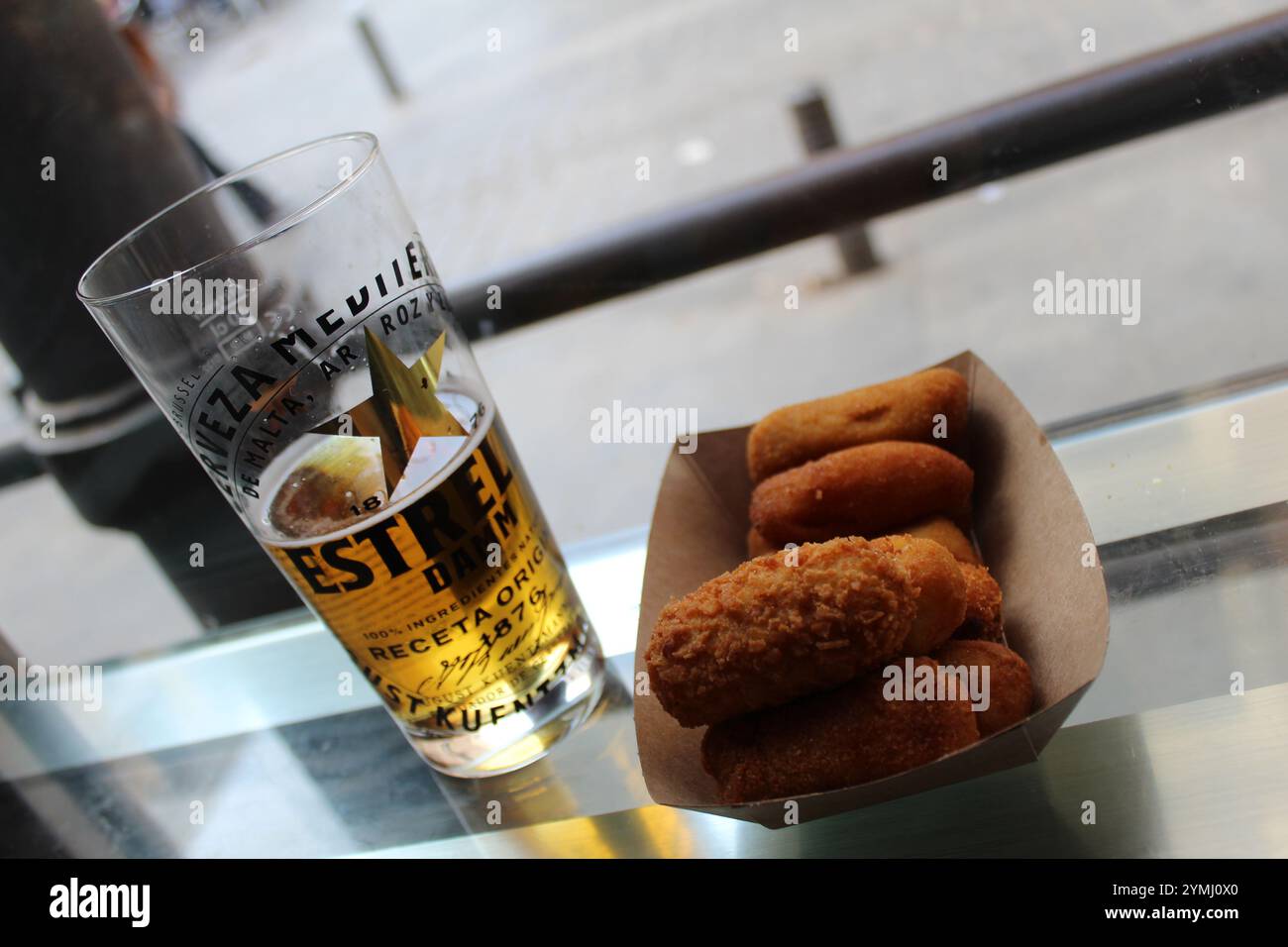 Bicchiere di birra Estrella Damm accompagnato da crocchette dorate e croccanti, collocate su un moderno tavolo di vetro in un ambiente urbano informale. Cultura gastronomica spagnola. Foto Stock