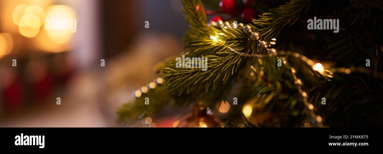 Ramo dell'albero di Natale adornato da luci a corda che creano un'atmosfera calda per le vacanze. Sfondo sfocato con decorazioni festose che esaltano l'atmosfera intima Foto Stock