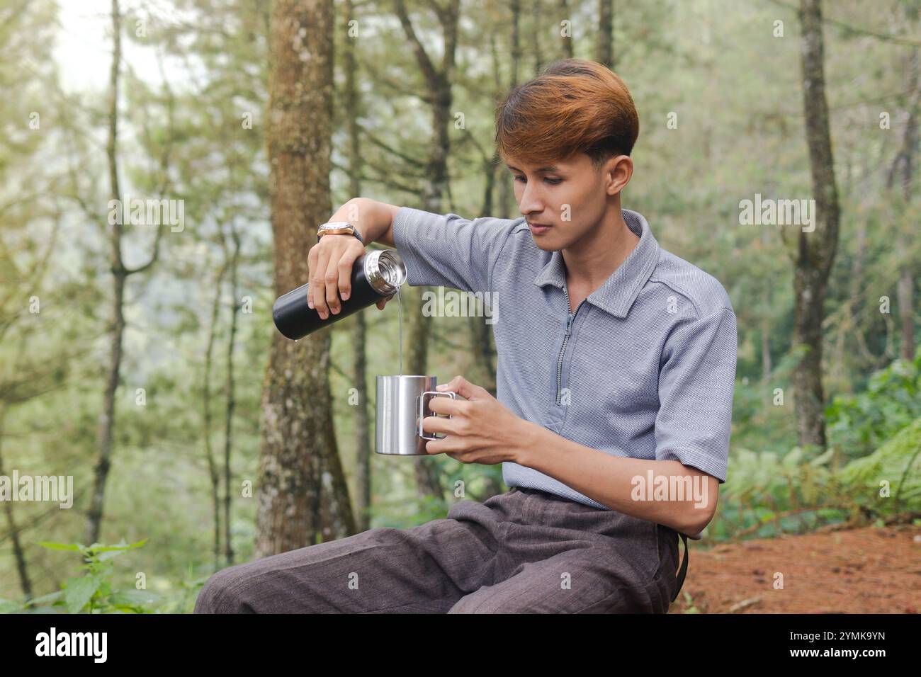 Ritratto di un uomo asiatico eccitato che versa acqua dalla fiasca del vuoto alla tazza in acciaio inossidabile. Bere acqua minerale durante il trekking nella foresta naturale Foto Stock