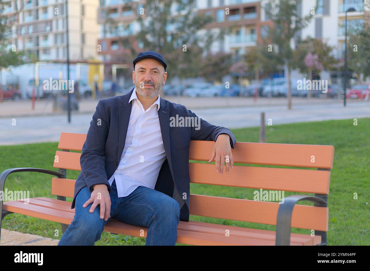 Uomo seduto su una panchina nel parco calmo e sorridente con un berretto ascot Foto Stock