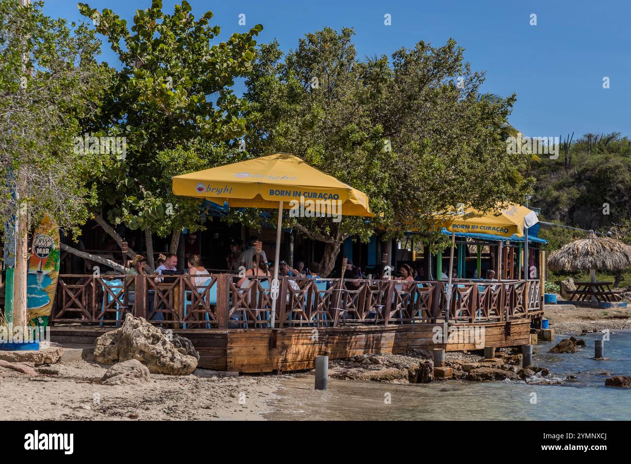 Il ristorante Brisa do Mar sulla spiaggia di Caracasbaaiweg, Jan Thiel, CuraCao, con una terrazza in legno proprio accanto all'acqua. Gli ombrelloni gialli recanti lo slogan «Born in CuraCao» fanno parte di una campagna pubblicitaria per il marchio di birra locale Brasa; questo popolare luogo di escursioni giornaliere si trova a circa dieci chilometri a est del centro di Willemstad. Kòrsou Foto Stock