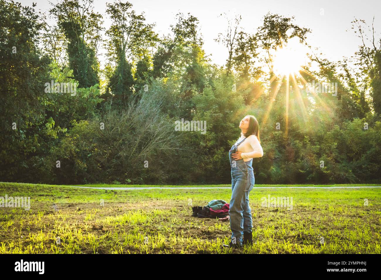 Contadino in piedi in un campo, godendosi la calda luce del sole che filtra tra gli alberi al tramonto Foto Stock