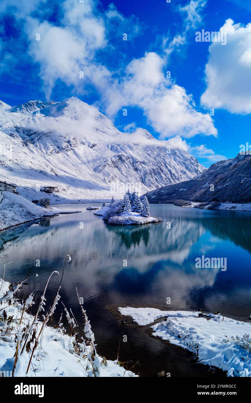 Una vista del pittoresco lago alpino con acqua blu scuro e riflessi, circondato da montagne innevate. In mezzo al lago - un piccolo Foto Stock