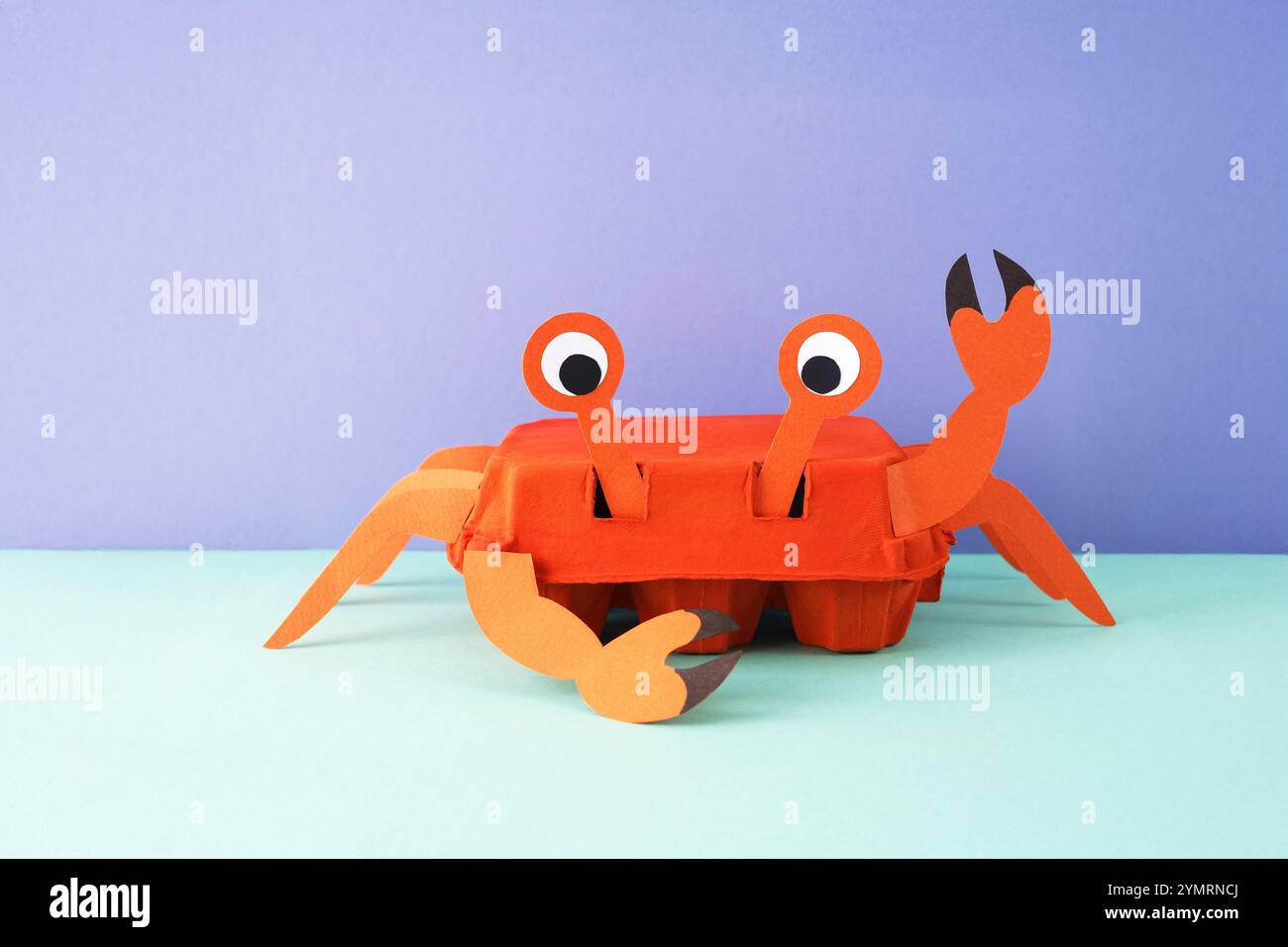 Crea un progetto che assomiglia al granchio realizzato in cartone d'uovo dipinto di arancione con gambe di carta, artigli e occhi. Artiglio ondulato e viso sorridente su viola chiaro A. Foto Stock