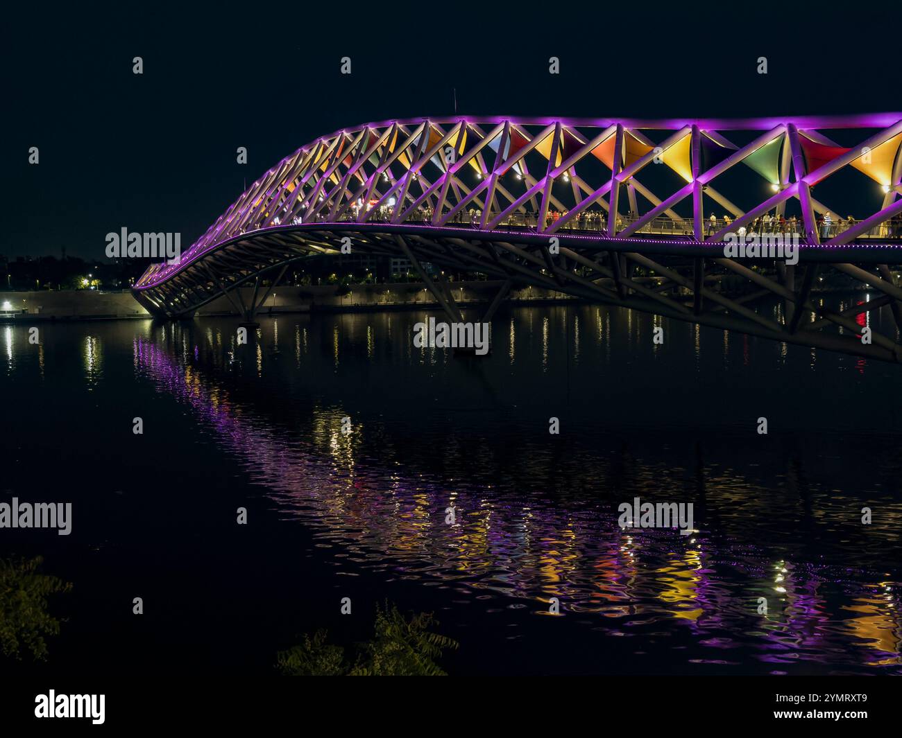Moderno ponte pedonale illuminato da vivaci luci viola che si riflettono sulle acque calme di notte Foto Stock