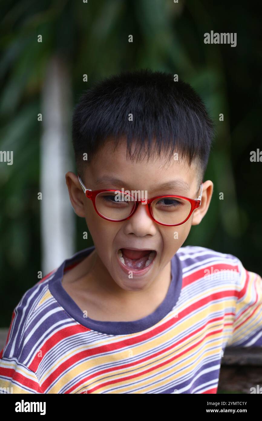 Un ragazzo gioioso con gli occhiali esprime emozioni all'aperto, mostrando energia ed emozioni in una camicia a righe casual circondata dal verde. Foto Stock
