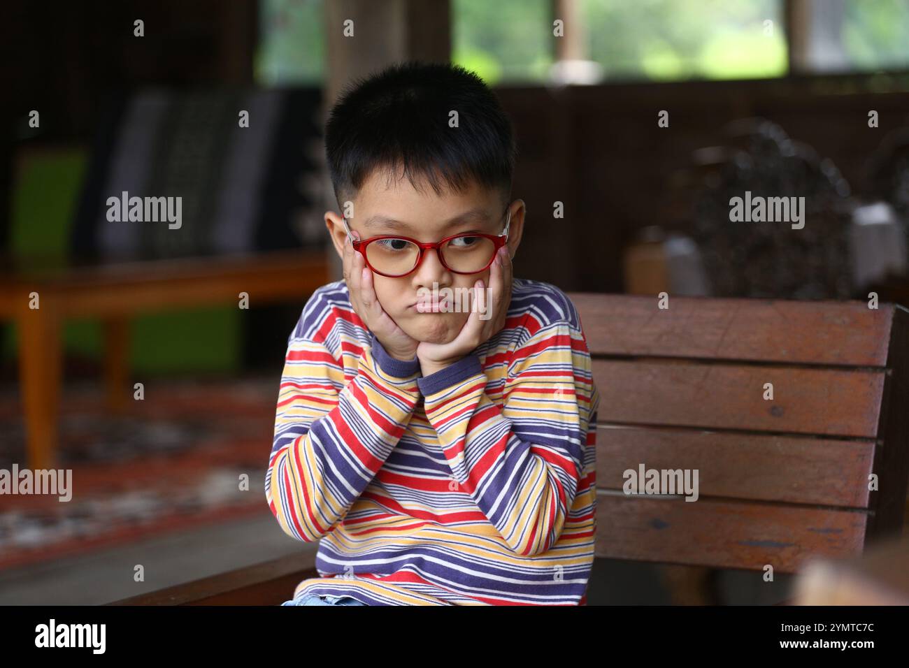 Un ragazzo premuroso con gli occhiali siede in casa, appoggiando il mento sulle mani. L'atmosfera intima crea uno sfondo perfetto per riflessi ed emozioni. Foto Stock