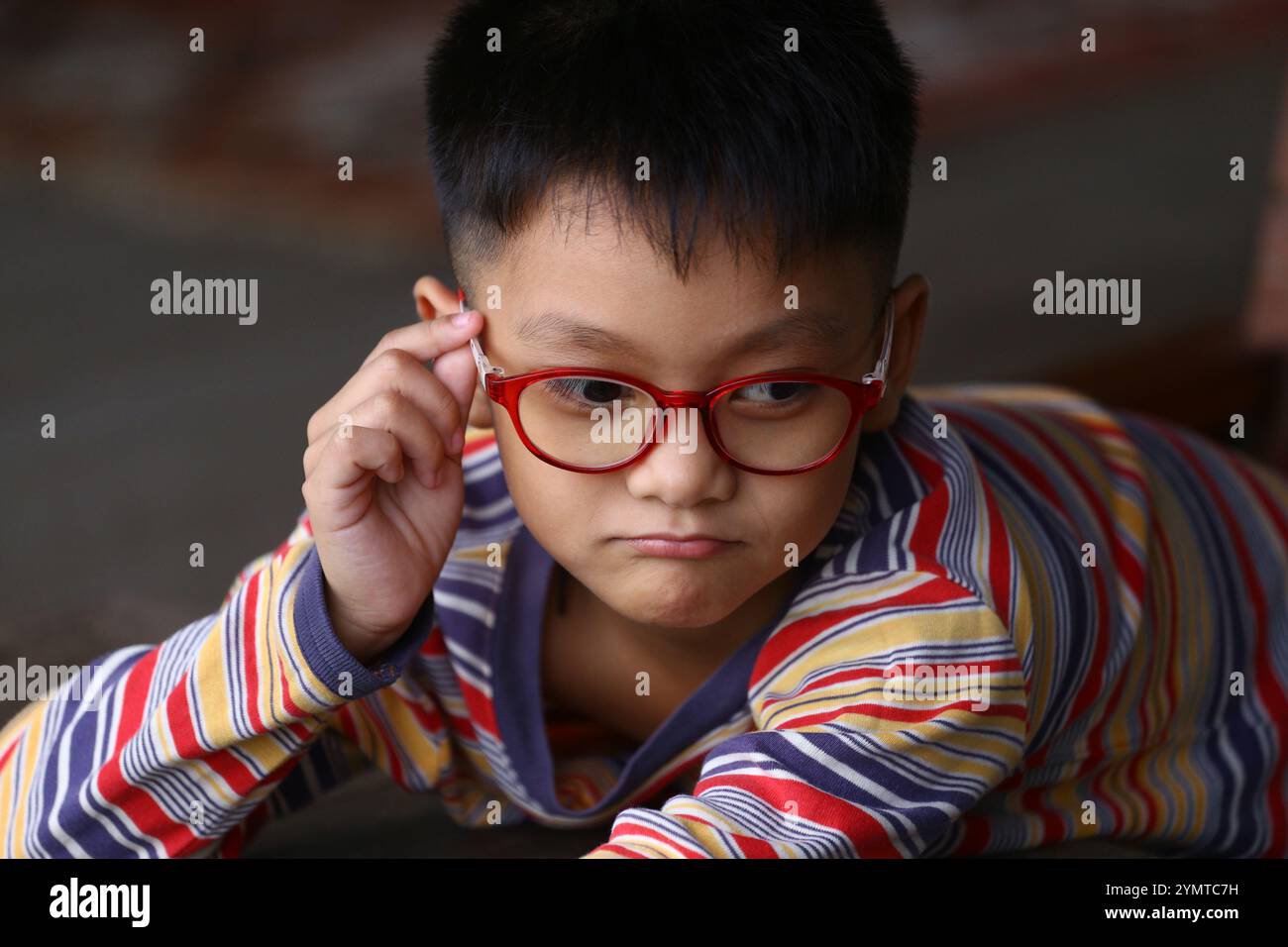Un ragazzo premuroso che indossa gli occhiali rossi ha un aspetto contemplativo mentre si sdraia sul pavimento in soffici pigiami. L'immagine cattura l'innocenza e la creatività in un i Foto Stock