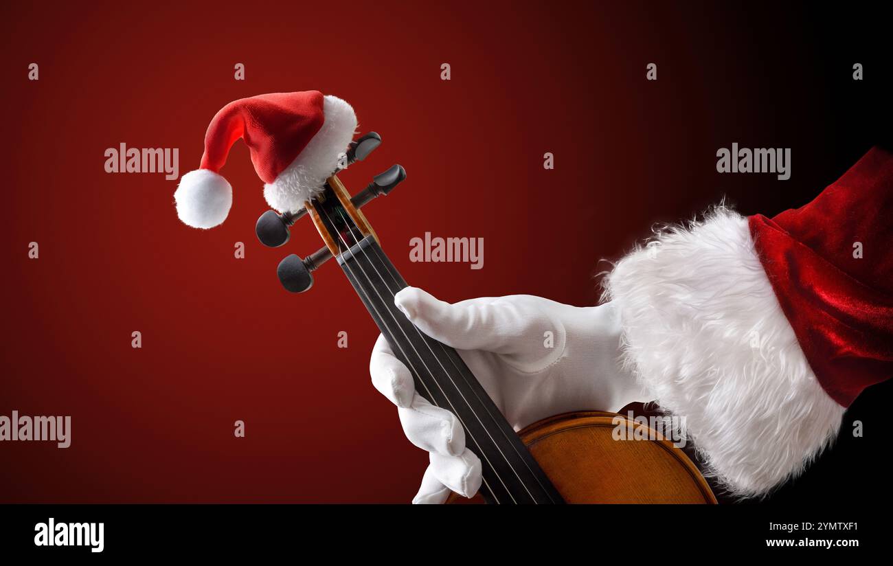 La mano di Babbo Natale regge un violino decorato con il cappello di Babbo Natale su uno sfondo isolato di sfumatura rossa. Vista frontale Foto Stock