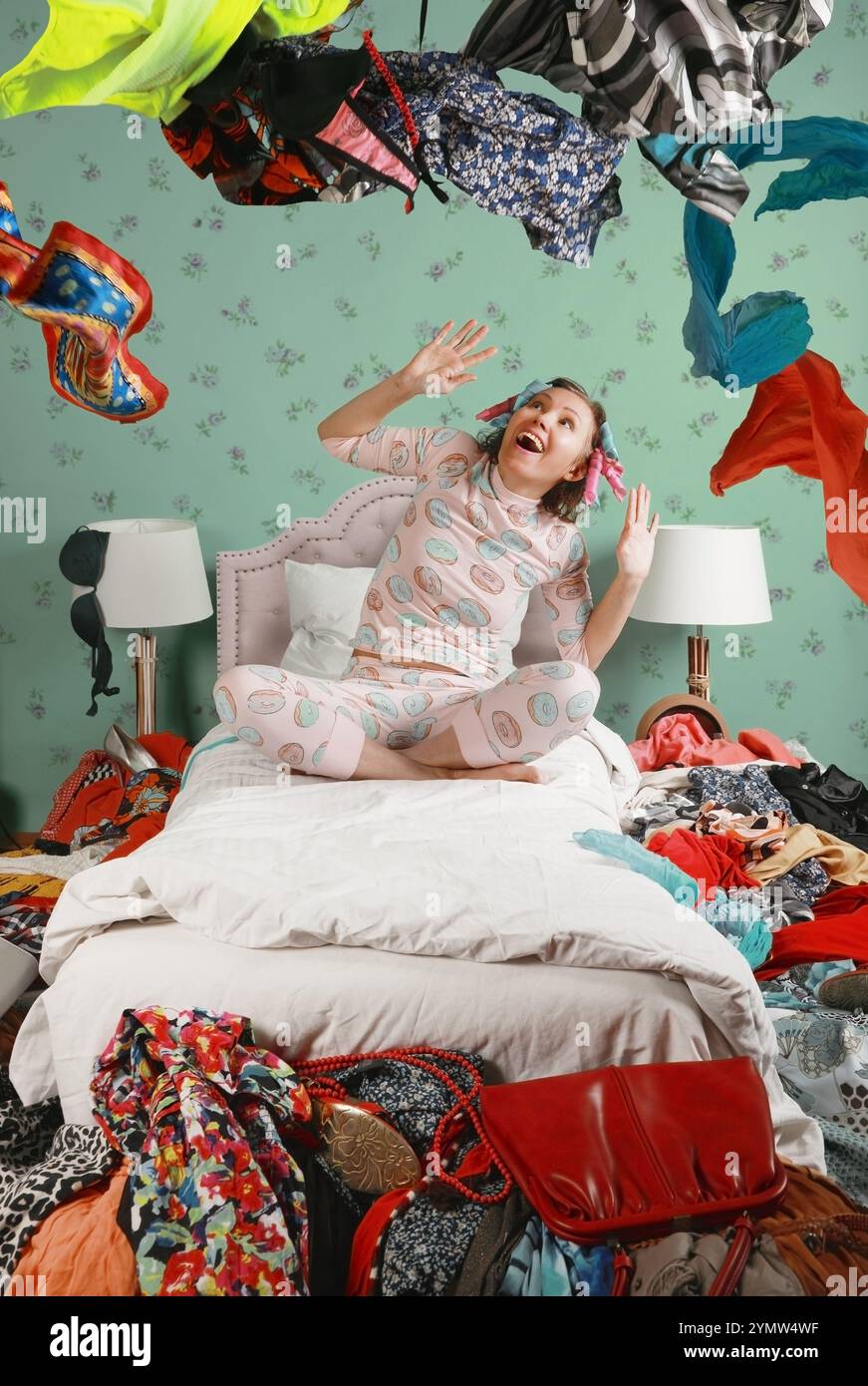Una donna in pigiama e bigiotteria si diverte a sedersi sul letto gettando cose in tutta la camera da letto. Il tema dello shopping e del consumo eccessivo di Foto Stock