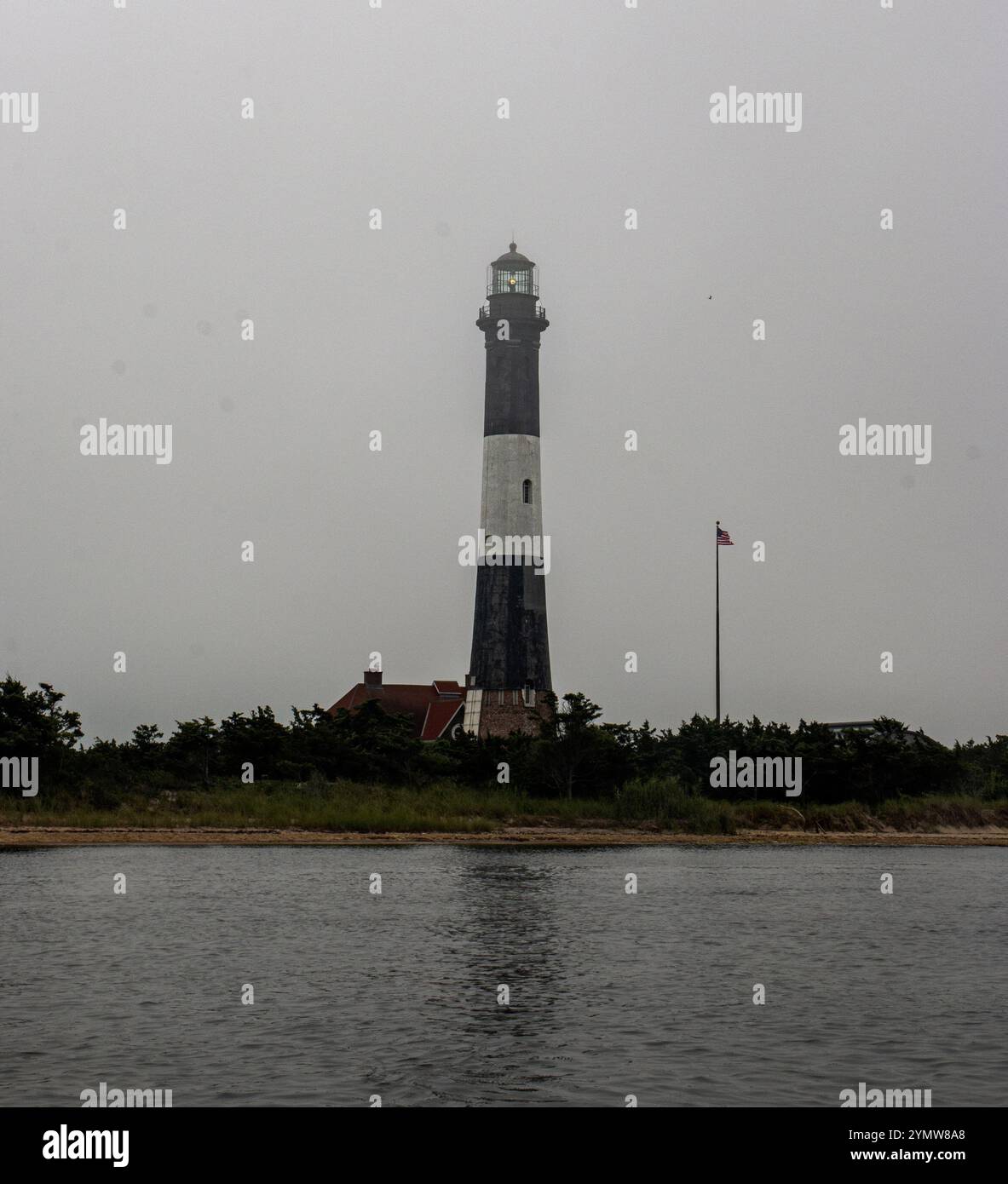 Un alto faro sorge sopra il bordo dell'acqua, circondato dal verde sotto un'atmosfera nuvolosa. Foto Stock