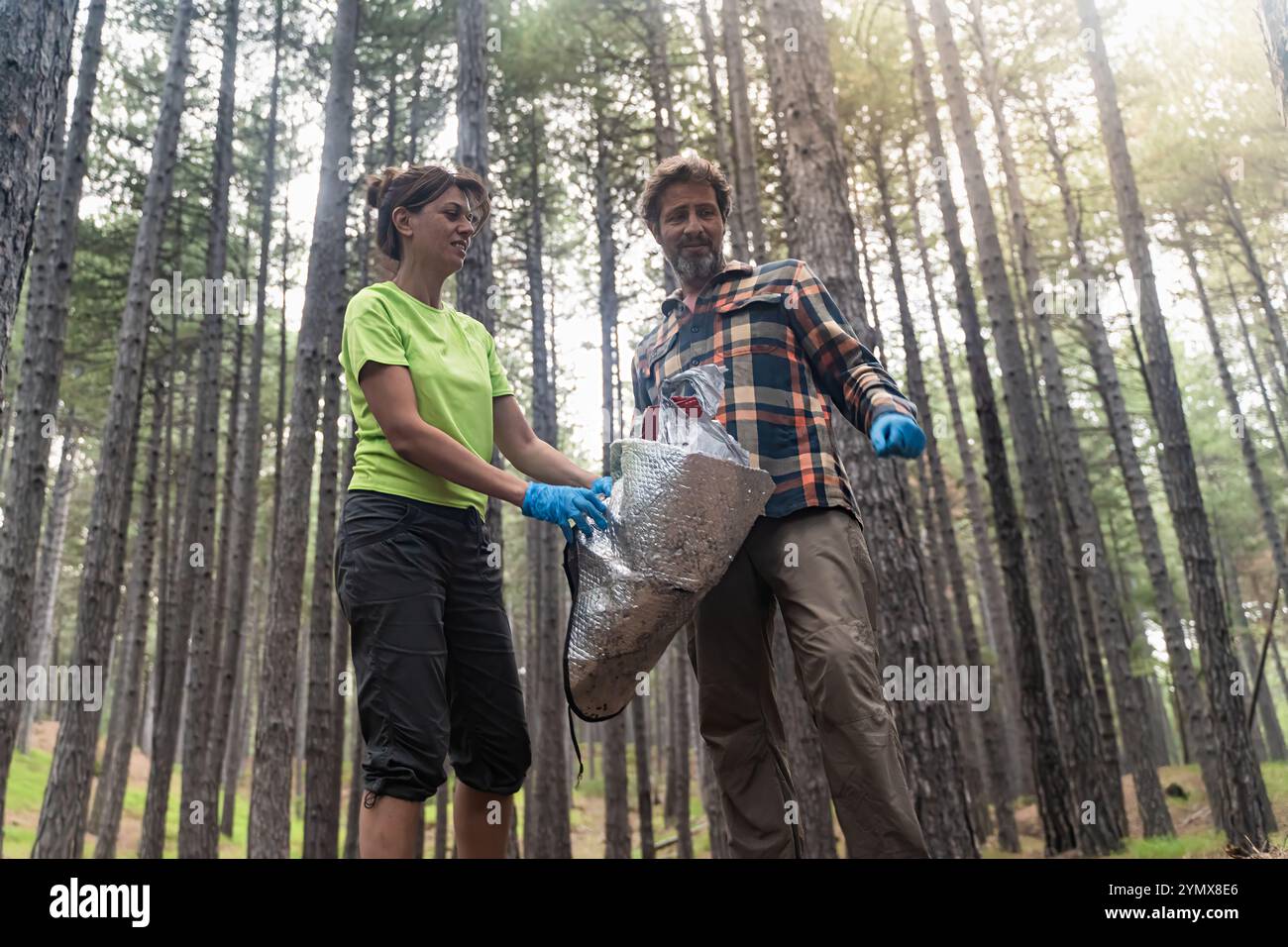 Allegri volontari sorridono mentre tengono i rifiuti raccolti nella foresta di pini. La vista ad angolo basso mostra un atteggiamento positivo durante la pulizia ambientale con il sole Foto Stock