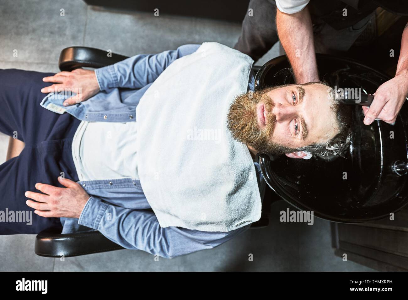 Uomo dai capelli di zenzero che si lava i capelli. Parrucchiere maschile che lava i capelli con prodotti per la pulizia profonda. Bel cliente che si rilassa con un asciugamano sul petto. Foto Stock