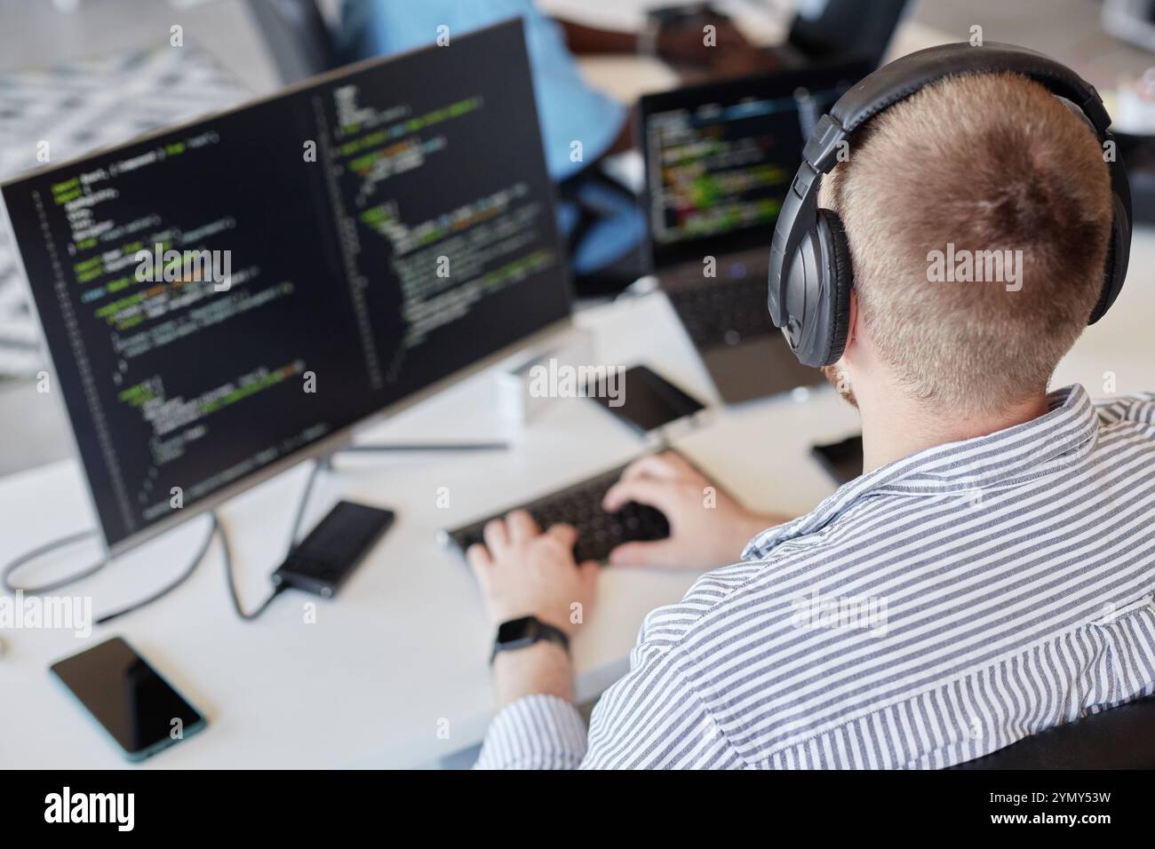 Uomo che indossa le cuffie seduto alla scrivania e che lavora al progetto di codifica su due monitor. L'immagine mostra l'ambiente di lavoro con vari dispositivi e strumenti surroun Foto Stock