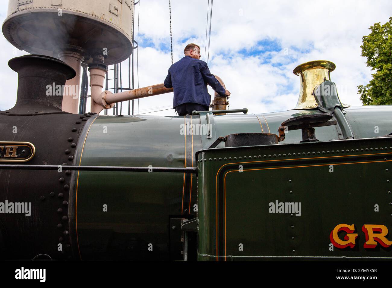 Un operaio si trova su una locomotiva d'epoca a vapore che regola l'approvvigionamento idrico, mostrando trasporti e ingegneria storici Foto Stock