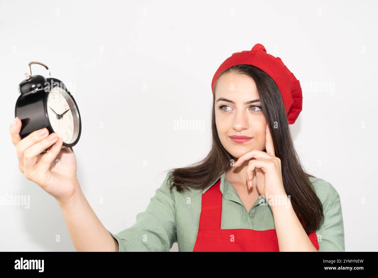 Giovane e premurosa casalinga in grembiule, cuoca fornaio che tiene l'orologio e pensa Foto Stock