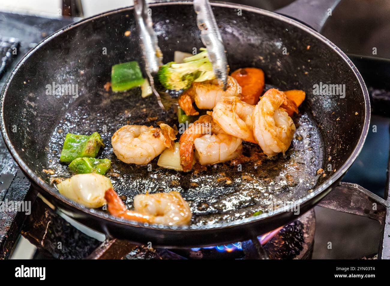 Gamberetti con peperoni, broccoli e carote sfrigolano in padella su una fiamma a gas aperta – uno dei piatti appena preparati a Plasa Bieu, la sala del mercato su De Ruyterkade a Willemstad, Curaçao. Le cucine aperte offrono agli ospiti una vista diretta della preparazione del cibo. Plasa Bieu è un mercato arioso e rustico con tavoli da picnic e cucine aperte che servono piatti classici di Curaçao. Foto Stock
