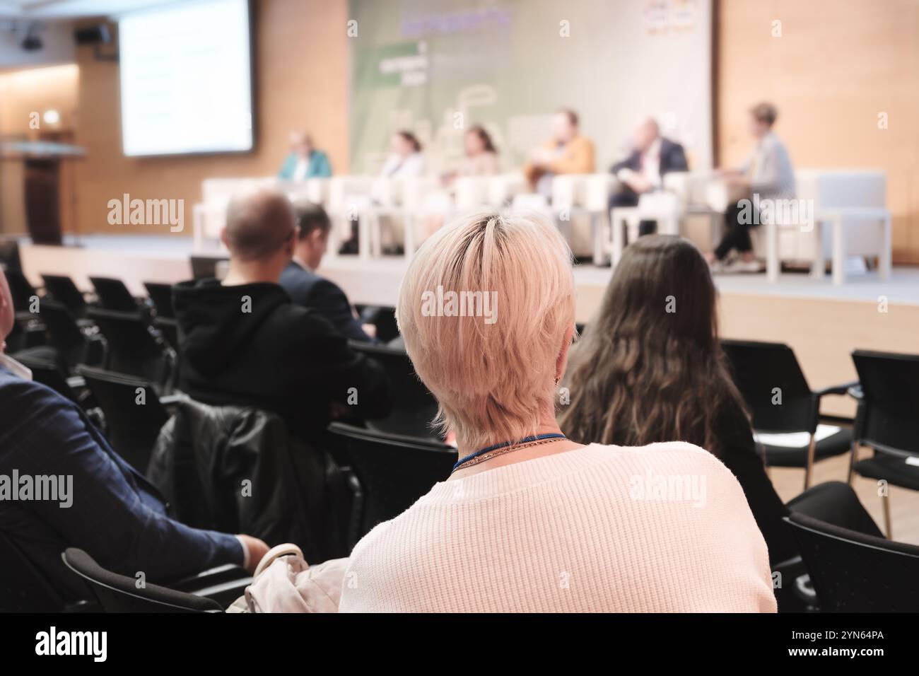 Una donna con una camicia bianca siede su una sedia a una conferenza. Ci sono molte altre persone nella stanza, alcune delle quali indossano anche camicie bianche Foto Stock