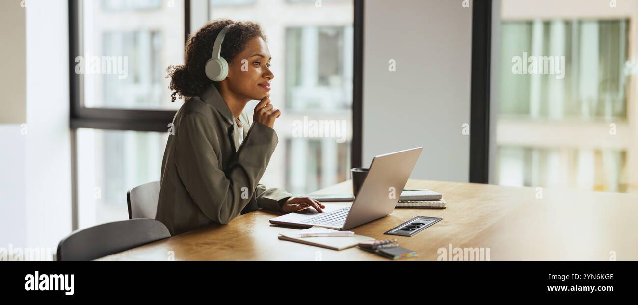 Una donna a un tavolo, che usa un computer portatile con le cuffie, vicino alla finestra di un edificio in un moderno ufficio di coworking Foto Stock