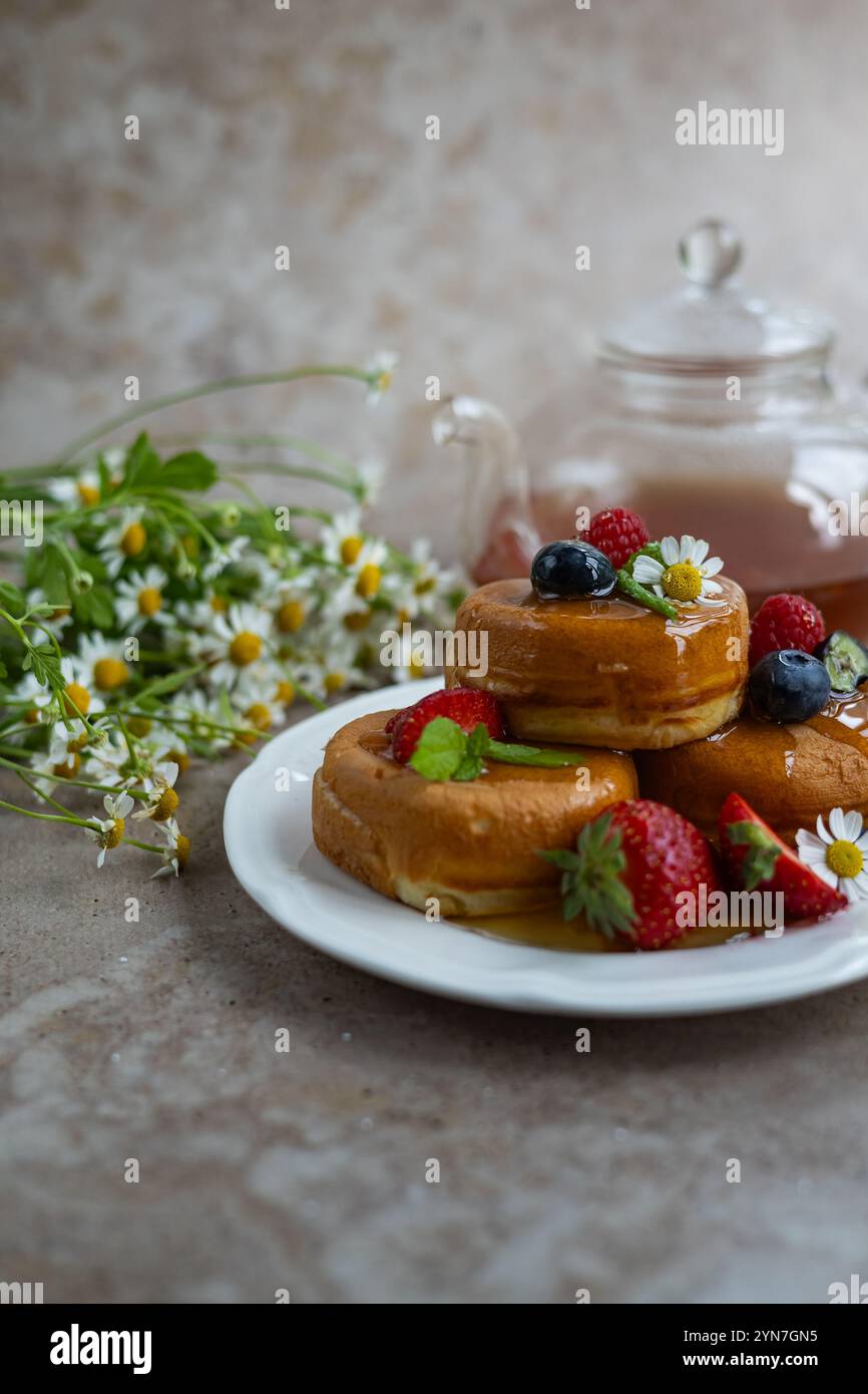 Soffici pancake giapponesi serviti con bacche fresche, foglie di menta e sciroppo, accompagnati da fiori di camomilla e una teiera di tè in vetro Foto Stock