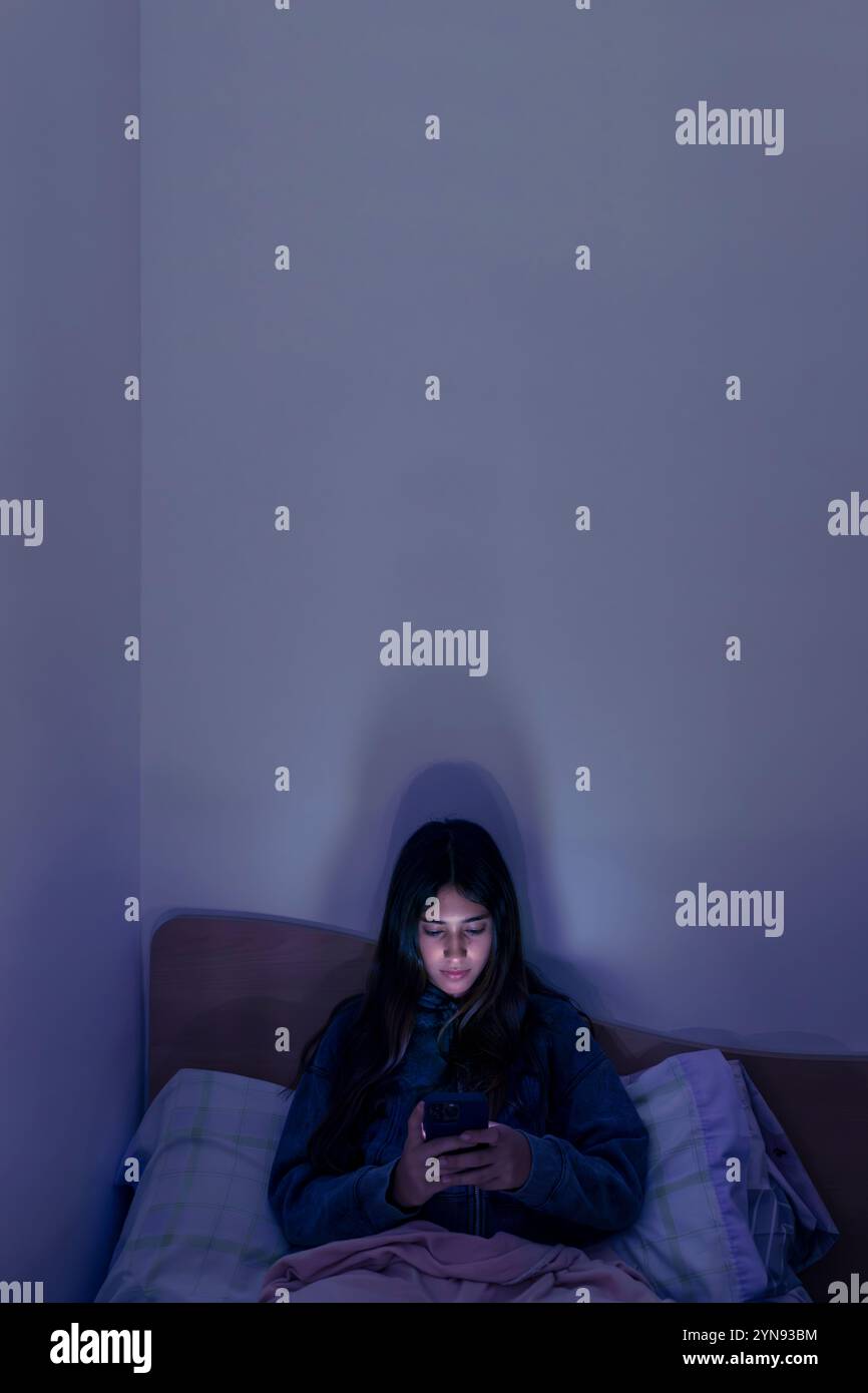 Immagine verticale di adolescente triste e stanca che utilizza uno smartphone sdraiato a letto a tarda notte, che gioca, guarda video e digita. Sc. Bambini Foto Stock