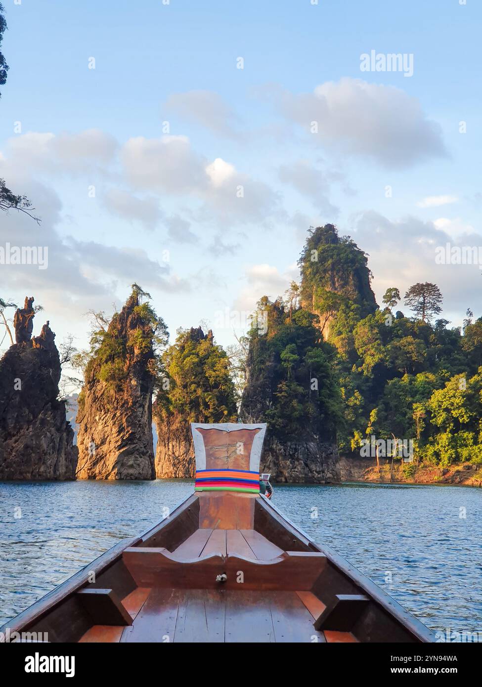 Scivolando sulle acque calme, una tradizionale barca in legno naviga tra le maestose scogliere calcaree bagnate dalla luce soffusa del mattino, mostrando la lussureggiante vegetazione della Thailandia Khao Sok Foto Stock