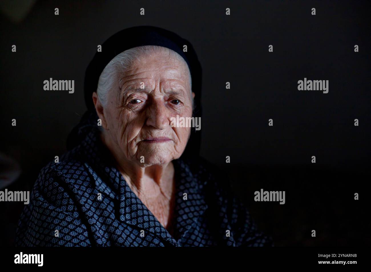 Una donna anziana che indossa un velo scuro siede tranquillamente in una stanza poco illuminata. La sua espressione rivela pensieri profondi, riflettendo una vita di esperienze e. Foto Stock