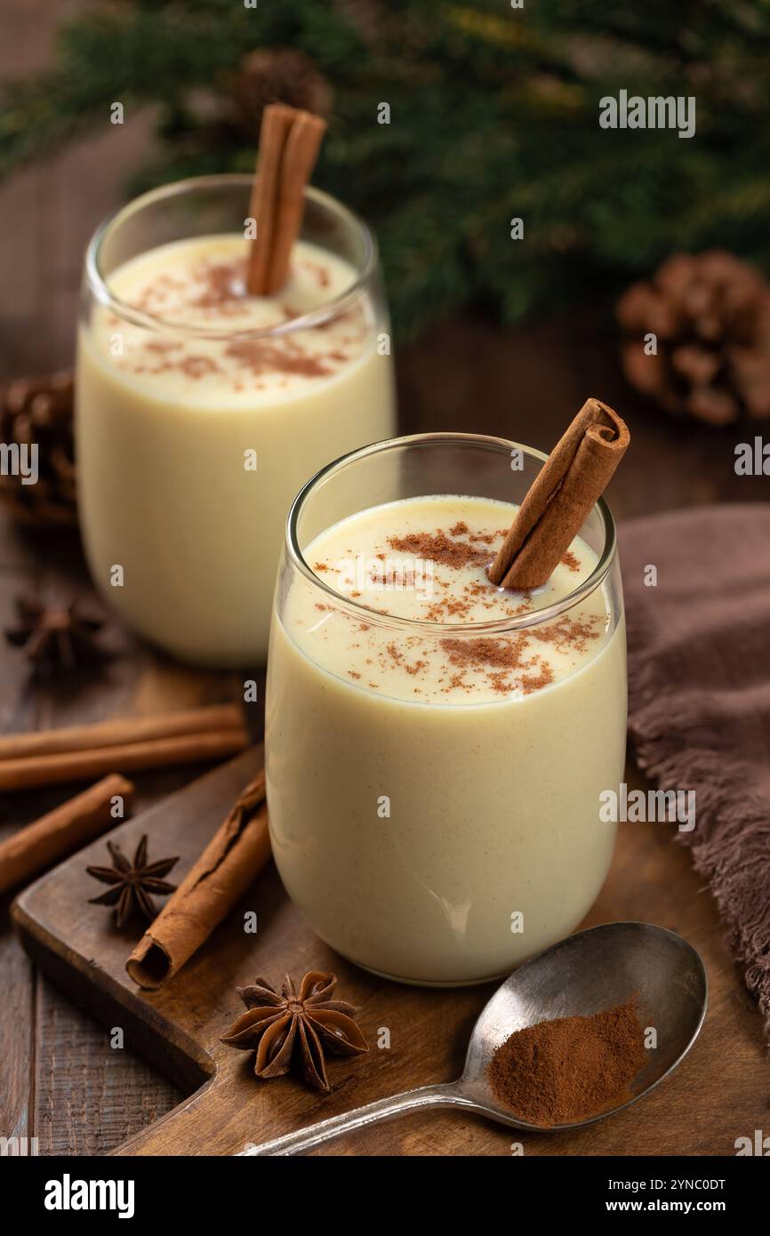 Due bicchieri di eggnog con cannella e anice su un tavolo di legno con decorazioni natalizie sullo sfondo Foto Stock